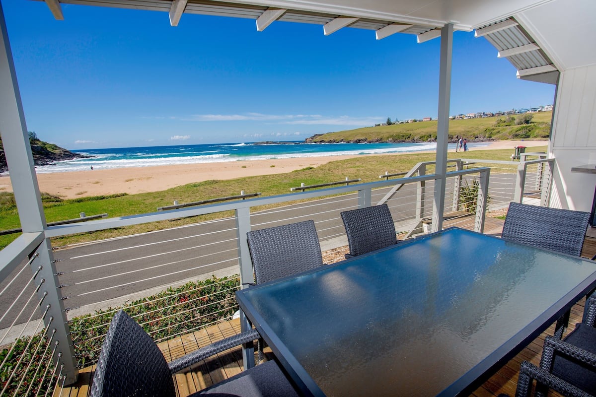 A covered outdoor seating area with a table and chairs, overlooking a beach and ocean.