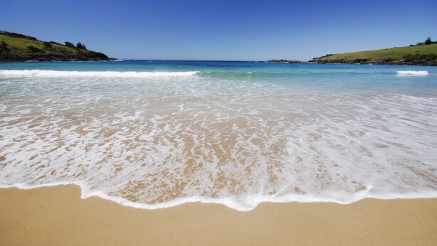 Uma praia com águas azul-claras, areal arenoso e colinas verdes ao fundo.