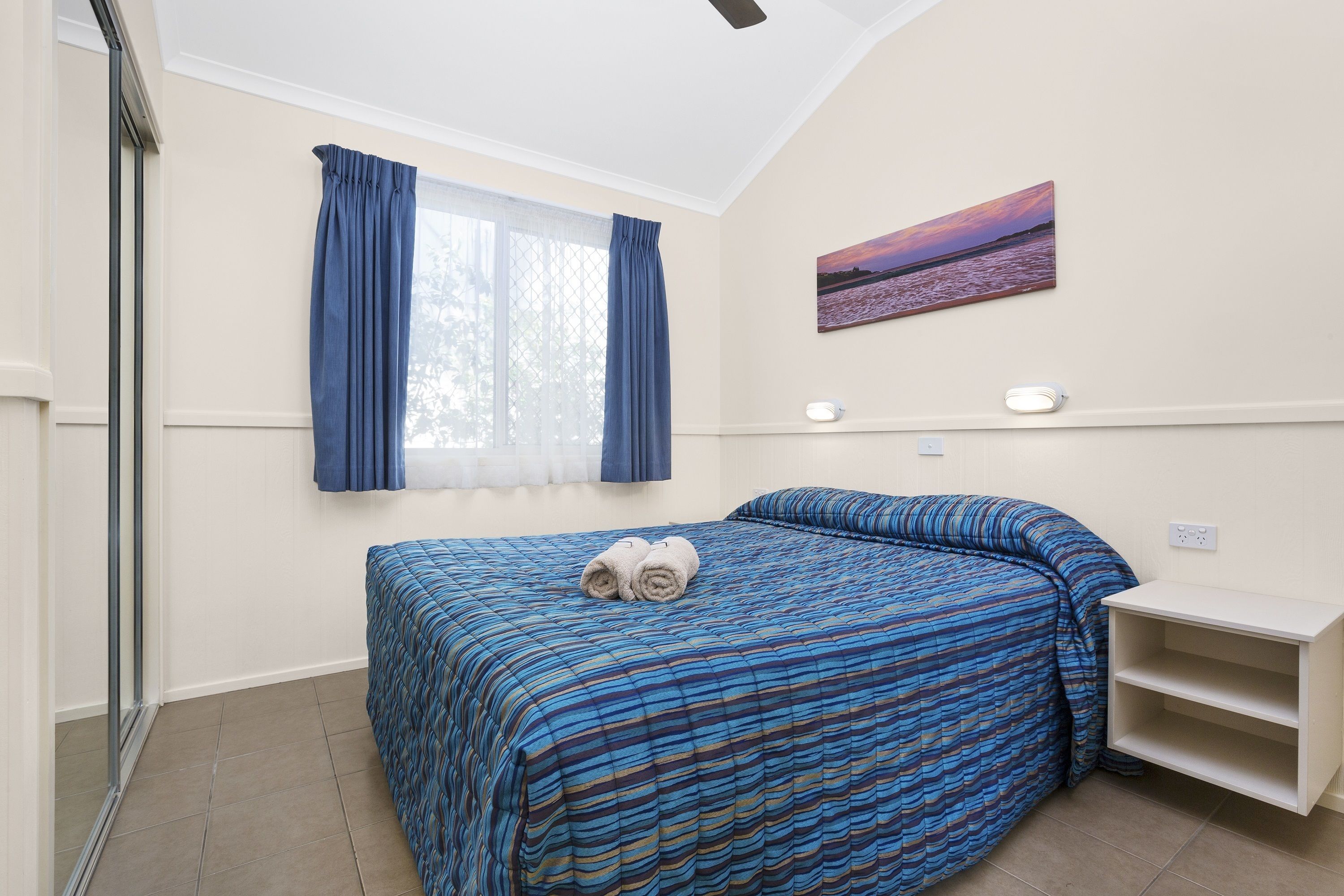 Un dormitorio con una cama, cortinas, un cuadro y una estantería.