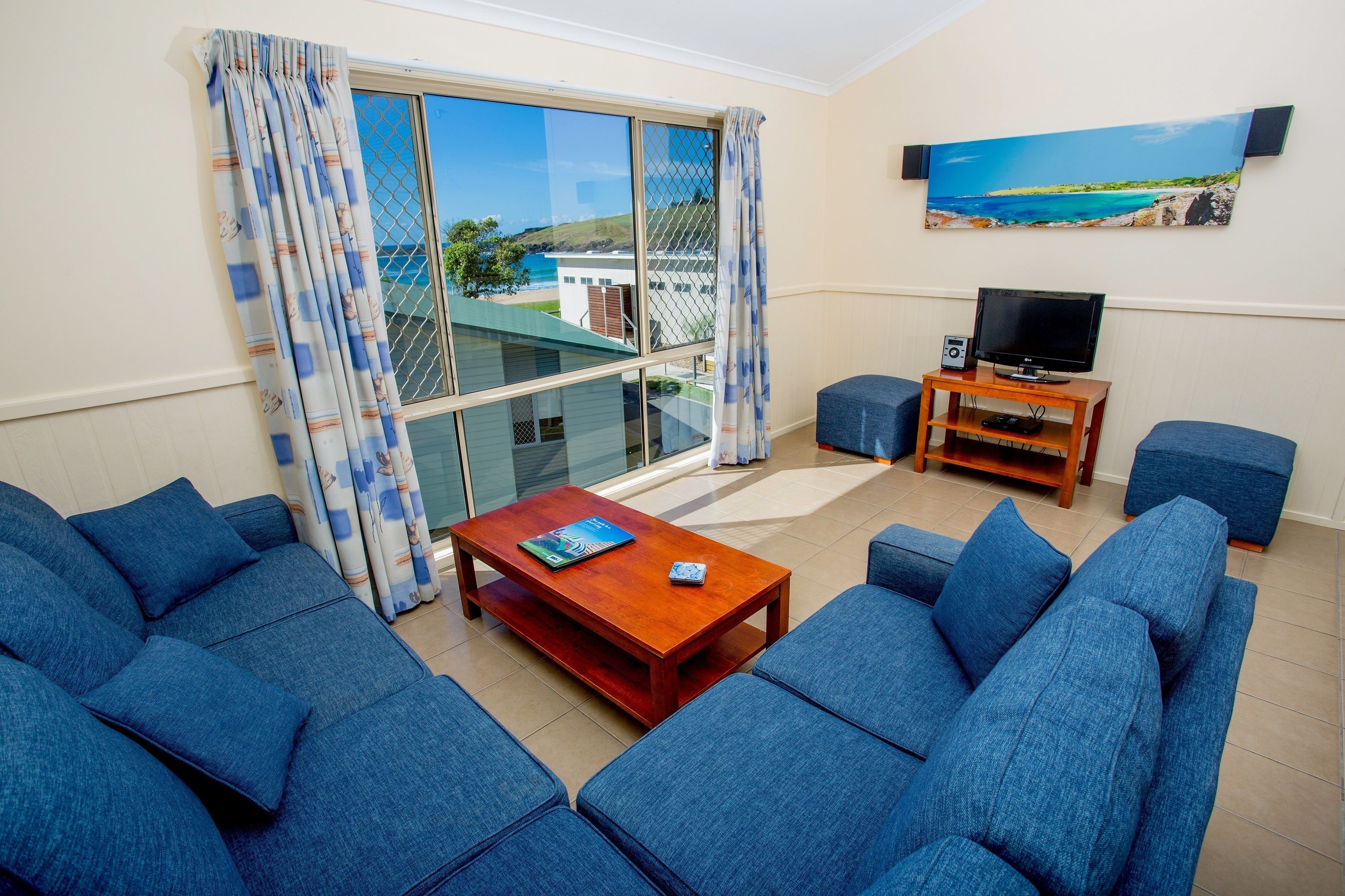 Ein Wohnzimmer mit blauen Sofas, einem Holztisch, einem Fernseher auf einem Holzschrank und einem großen Fenster mit Blick auf das Meer.