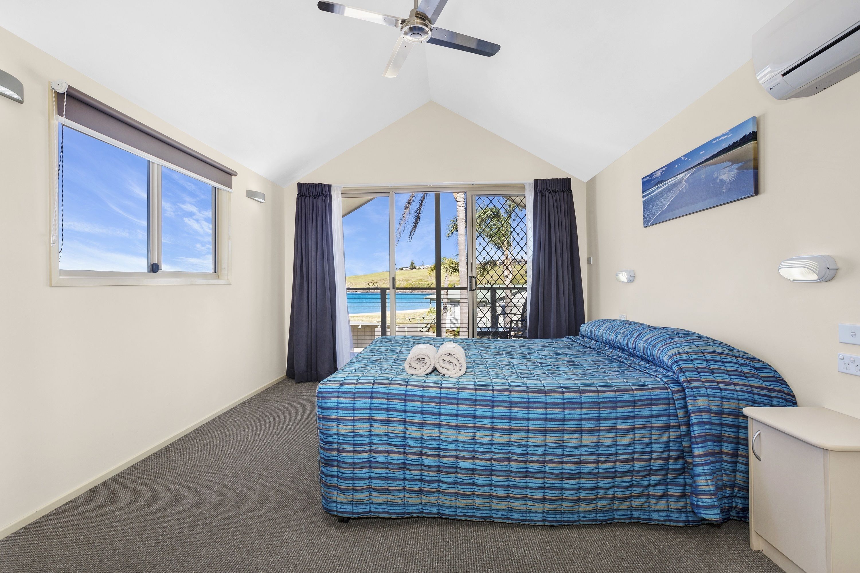 A bedroom with a large bed, a ceiling fan, a window with curtains, a view of the outdoors, and a painting on the wall.