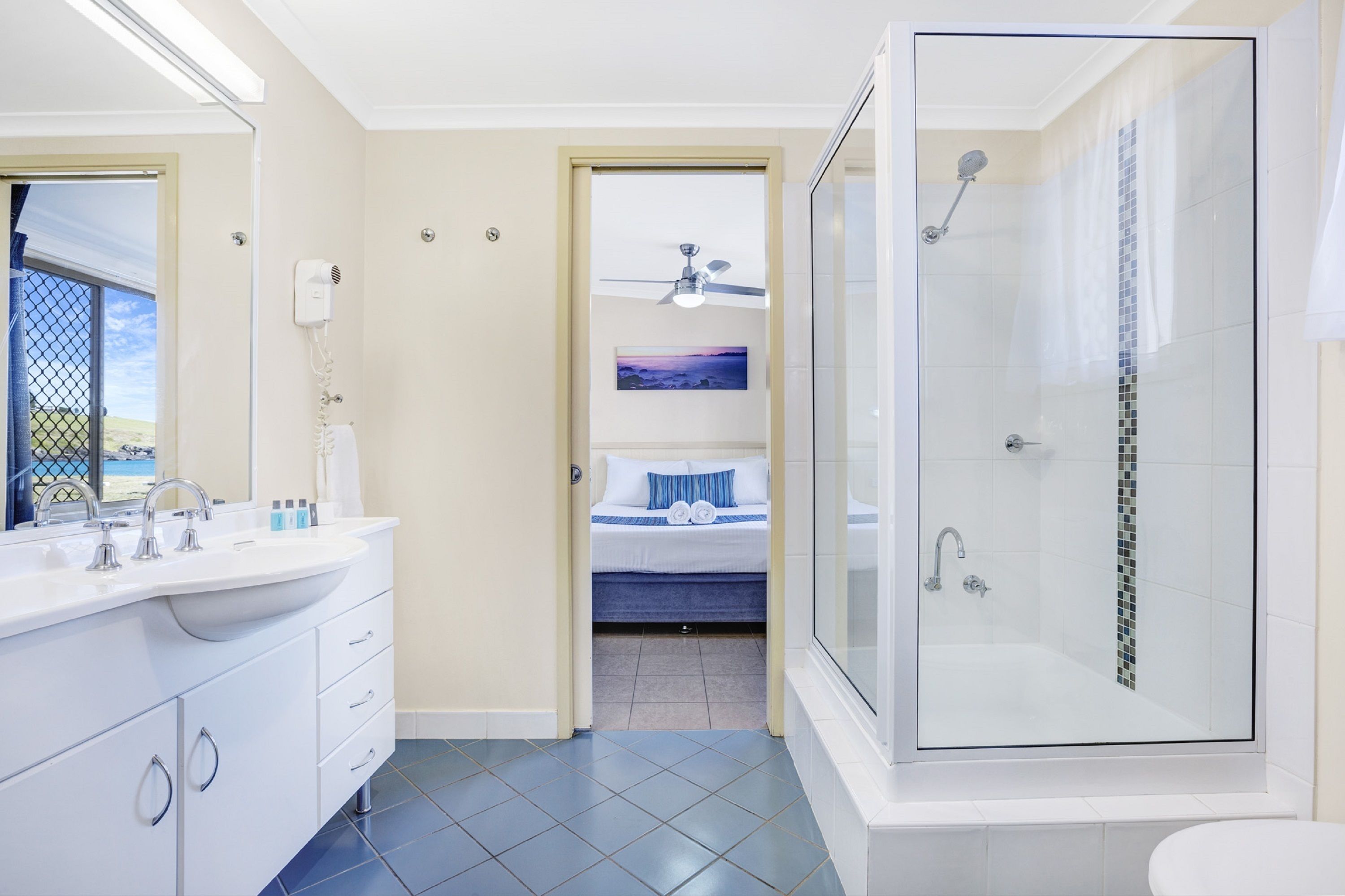 Un baño moderno con ducha de vidrio, un lavamanos grande con espejo y una cama con vista al atardecer.