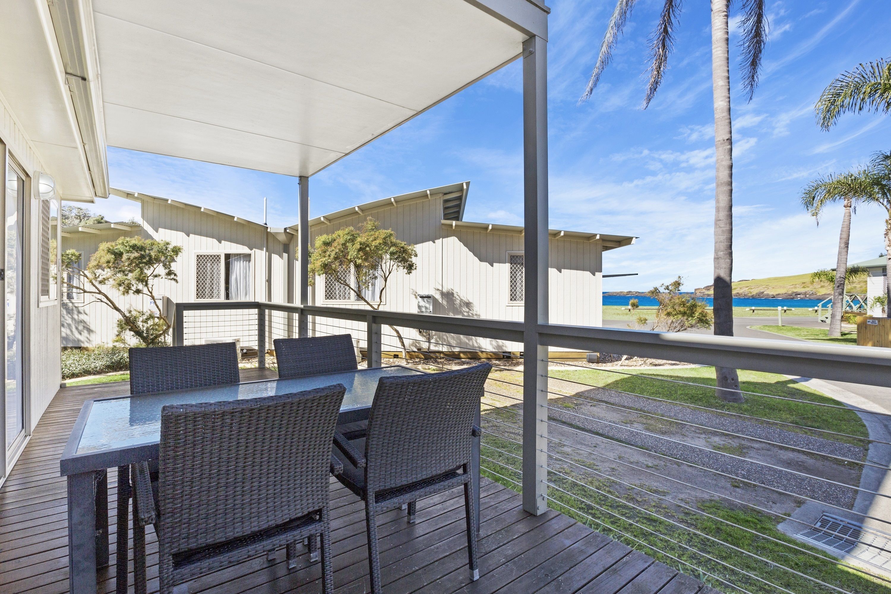 Een overdekt terras met een tafel en stoelen, met uitzicht op een strand en palmbomen.