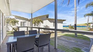 Een overdekt terras met een tafel en stoelen, met uitzicht op een strand en palmbomen.