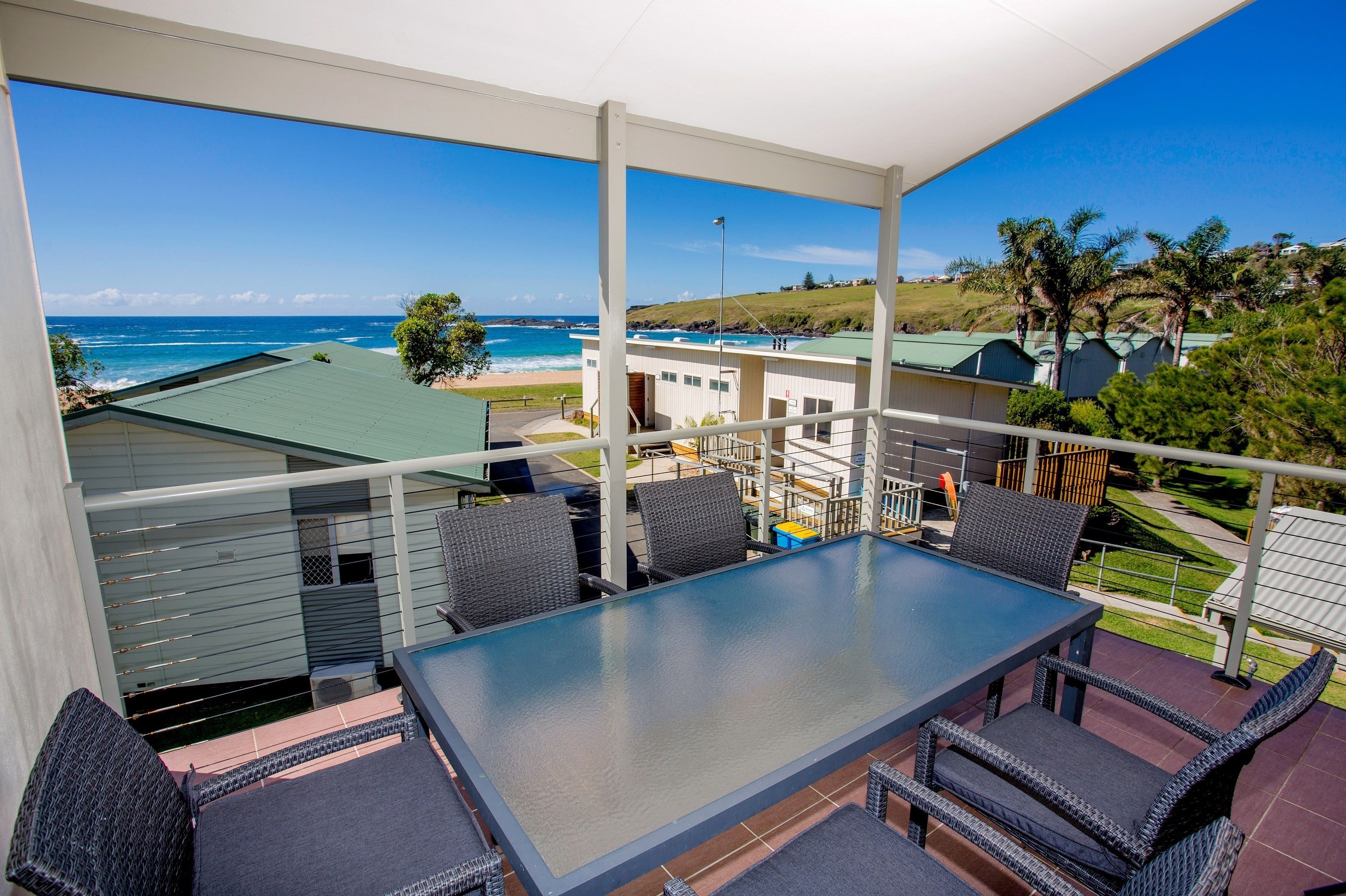 A balcony with a glass table and wicker chairs, overlooking a beach and residential buildings.