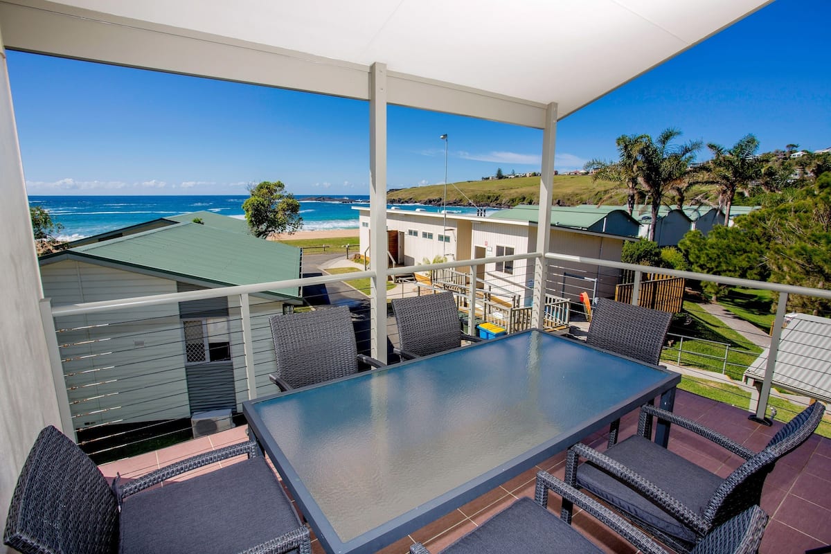 A balcony with a glass table and wicker chairs, overlooking a beach and residential buildings.
