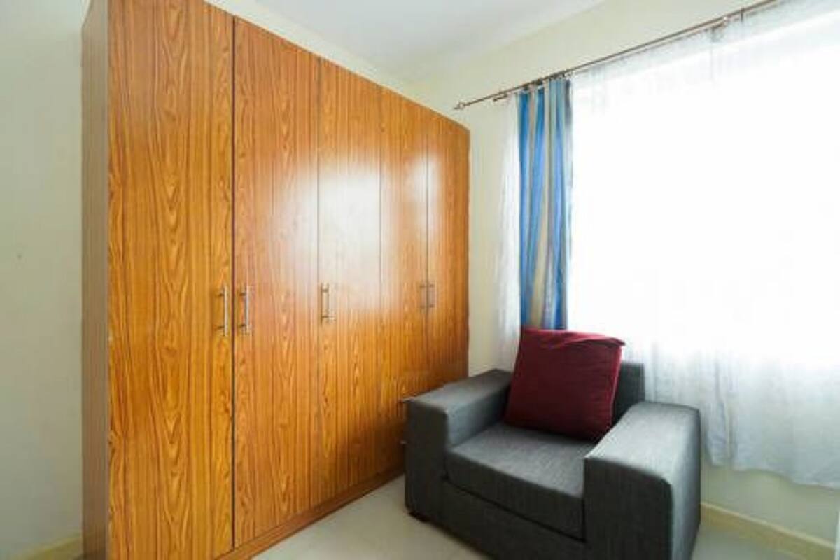 A room with a wooden wardrobe, a grey sofa with a red cushion, and a window with blue curtains.