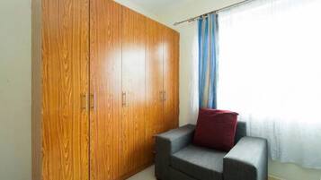 A room with a wooden wardrobe, a grey sofa with a red cushion, and a window with blue curtains.