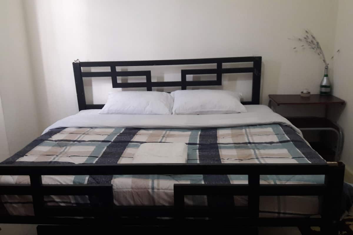 A single bed with a black metal frame, white bedding, and a plaid blanket. There is a small bedside table with a vase of flowers.