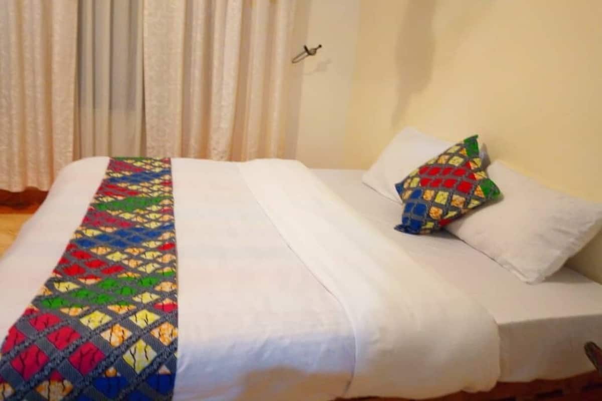 A bed with a colorful quilt and a single pillow in a room with a curtain and a wall-mounted hook.