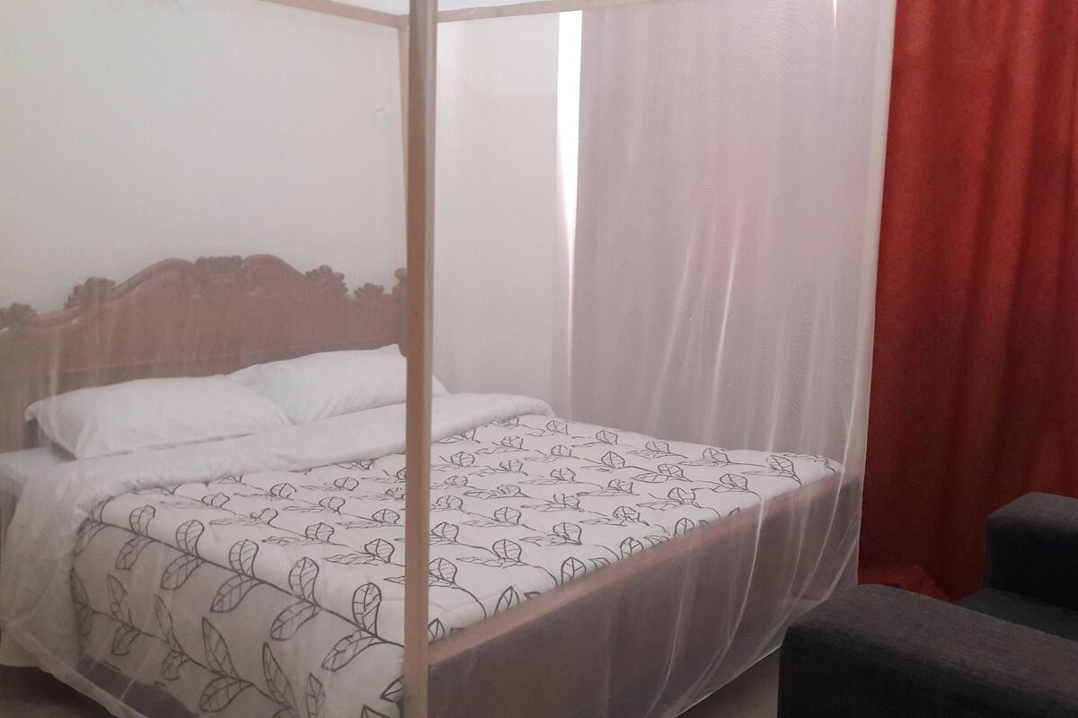 A room with a four-poster bed, a canopy, a red curtain, and a grey armchair.