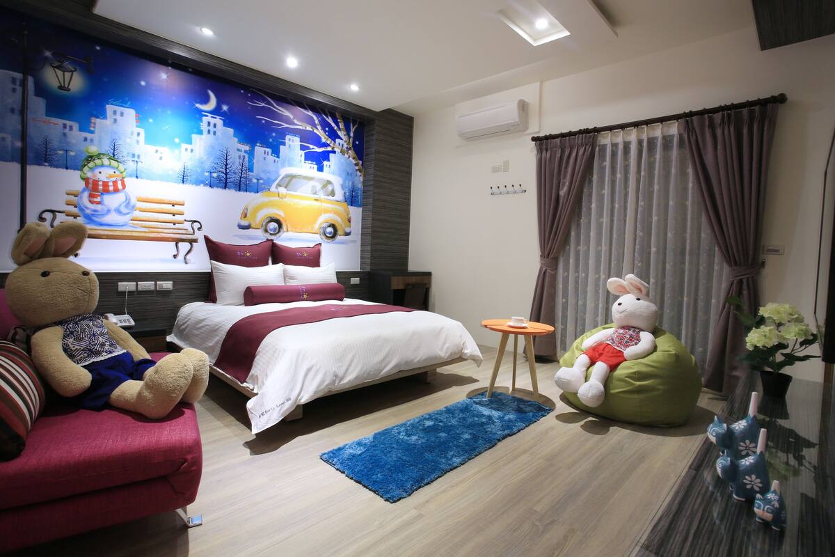 Habitación de hotel moderna con cama, sofá, sillón de peluche y un mural en la pared.