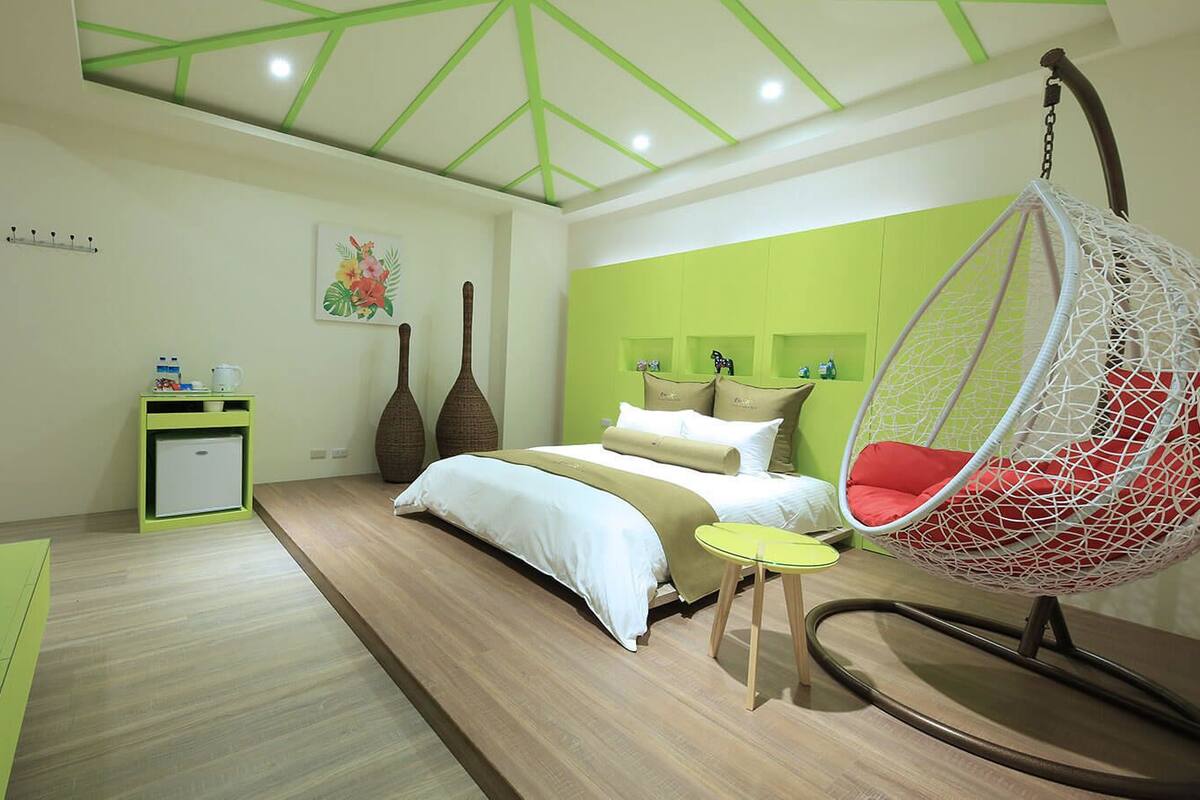 Un dormitorio moderno con una silla colgante, una cama con ropa de cama blanca, una mesita pequeña y una pared verde.