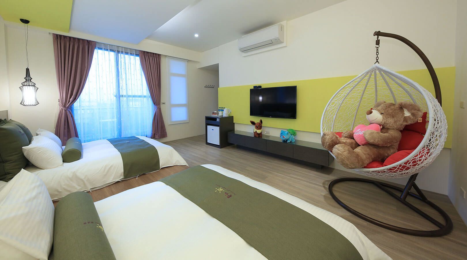 A hotel room with two beds, a television, a desk, and a hanging chair with a teddy bear.