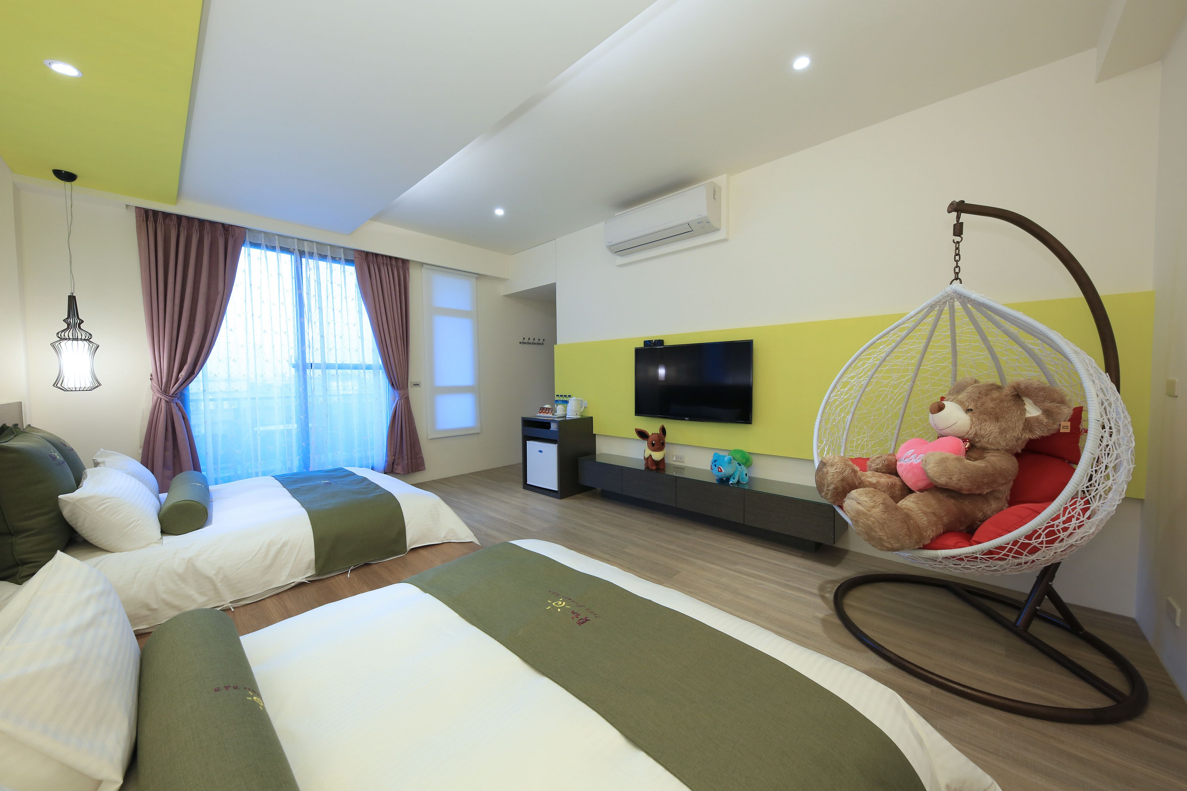 A hotel room with two beds, a hanging chair with a teddy bear, a TV on the wall, and a window with curtains.