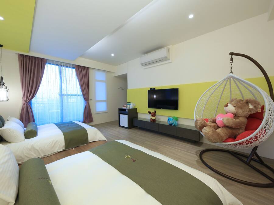 A hotel room with two beds, a hanging chair with a teddy bear, a TV on the wall, and a window with curtains.