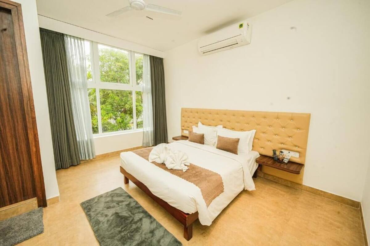 Quarto de hotel com uma cama grande, cabeceira de madeira, mesa de cabeceira e janela com cortinas.