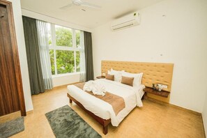 A hotel room with a large bed, a wooden headboard, a nightstand, and a window with curtains. - Shelter Beach Resort (Tirupporur)