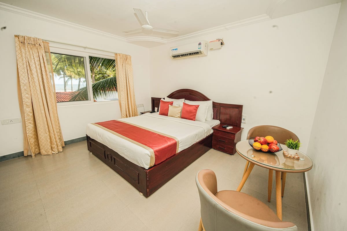 Quarto de hotel com cama, uma cadeira, uma pequena mesa com frutas, janela com cortinas e um ar condicionado.