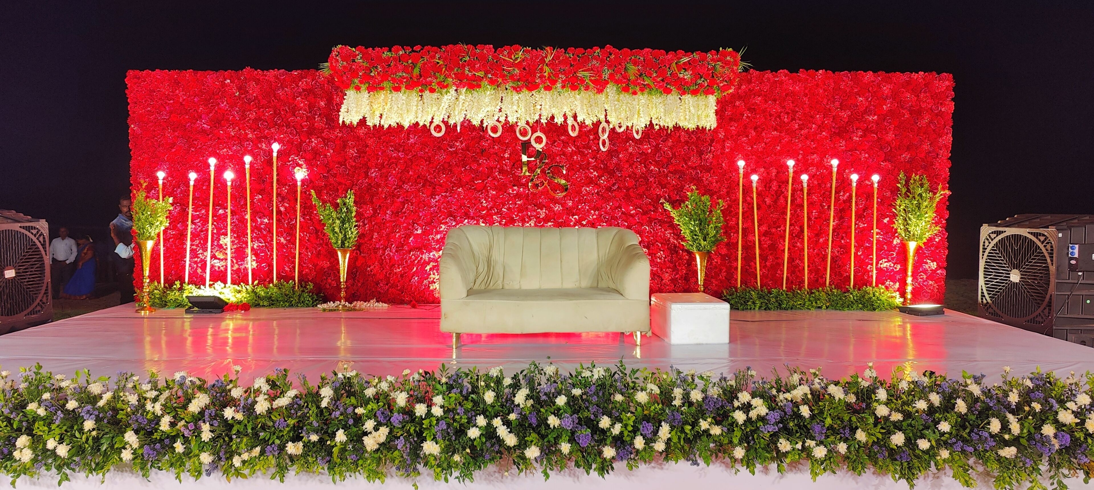 A stage with a sofa and a white table, surrounded by red floral decorations and a border of mixed flowers.
