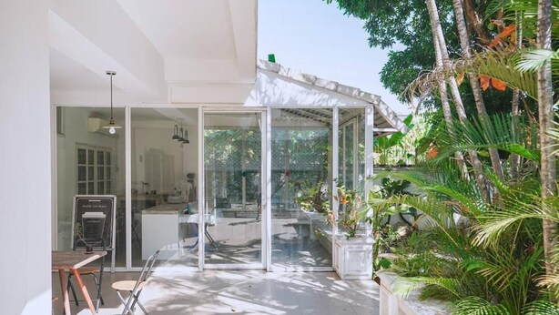 A modern outdoor space with a glass enclosure, a dining area with chairs, and lush tropical plants.