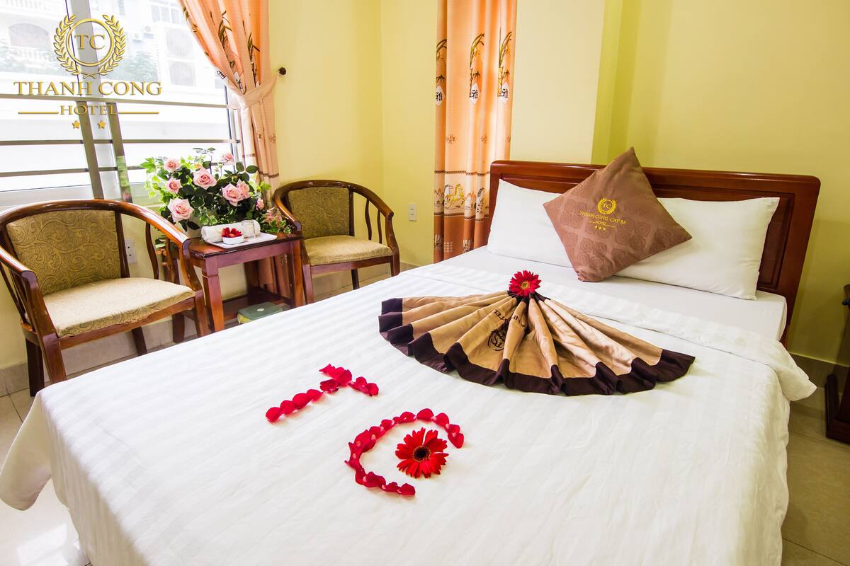 A hotel room with a bed, two chairs, a small table with a vase of flowers, and a window with curtains.