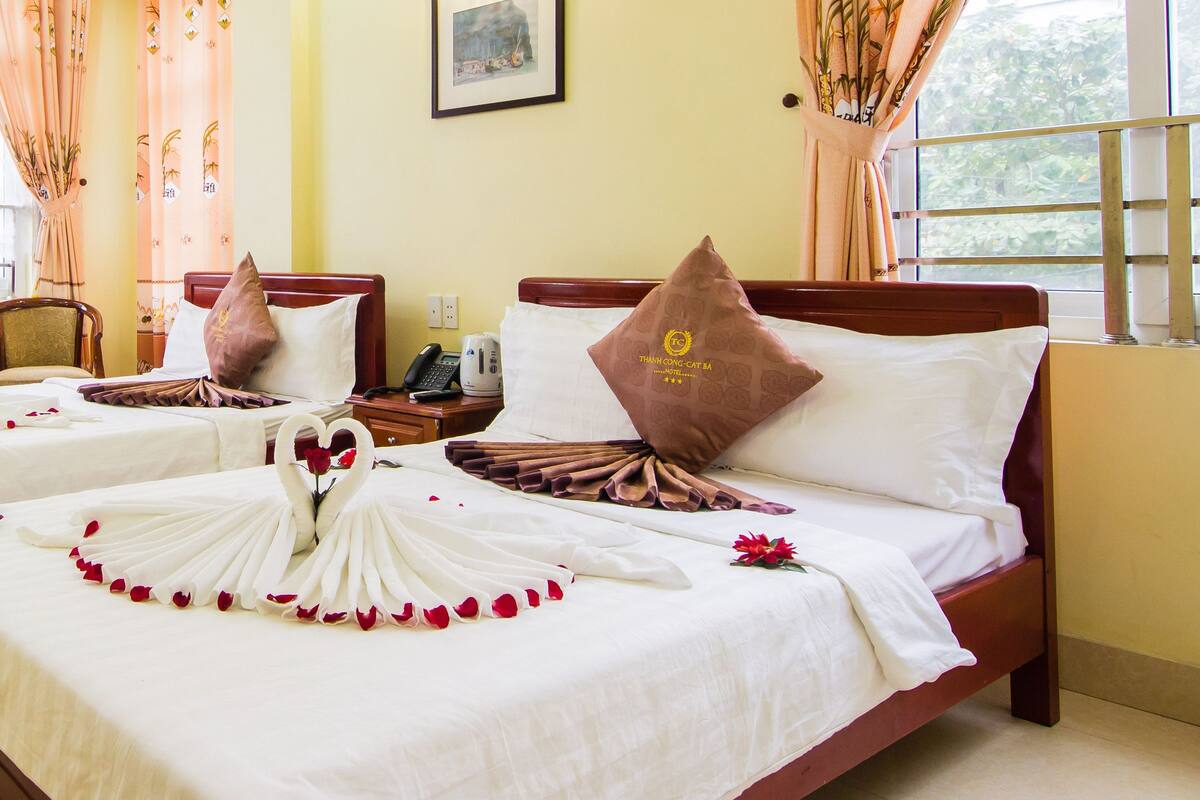 A hotel room with two beds, each with a swan-shaped arrangement of white and red rose petals. There are brown pillows with a logo, a framed picture on the wall, and a window with curtains.