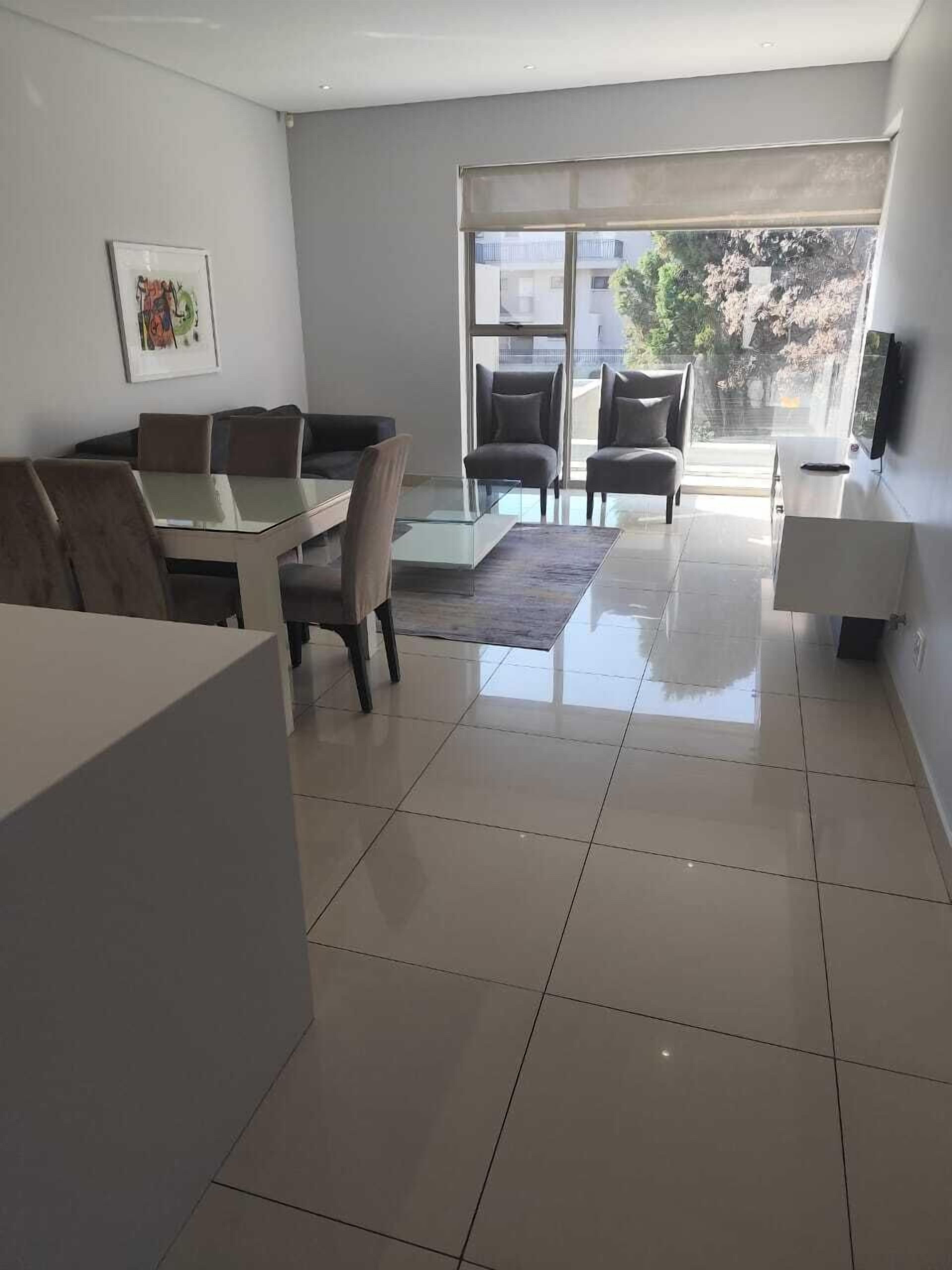 Una sala moderna con una mesa de comedor de vidrio, asientos cómodos, un televisor de pantalla plana y un ventanal con vistas al exterior.