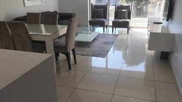 Una sala moderna con una mesa de comedor de vidrio, asientos cómodos, un televisor de pantalla plana y un ventanal con vistas al exterior.