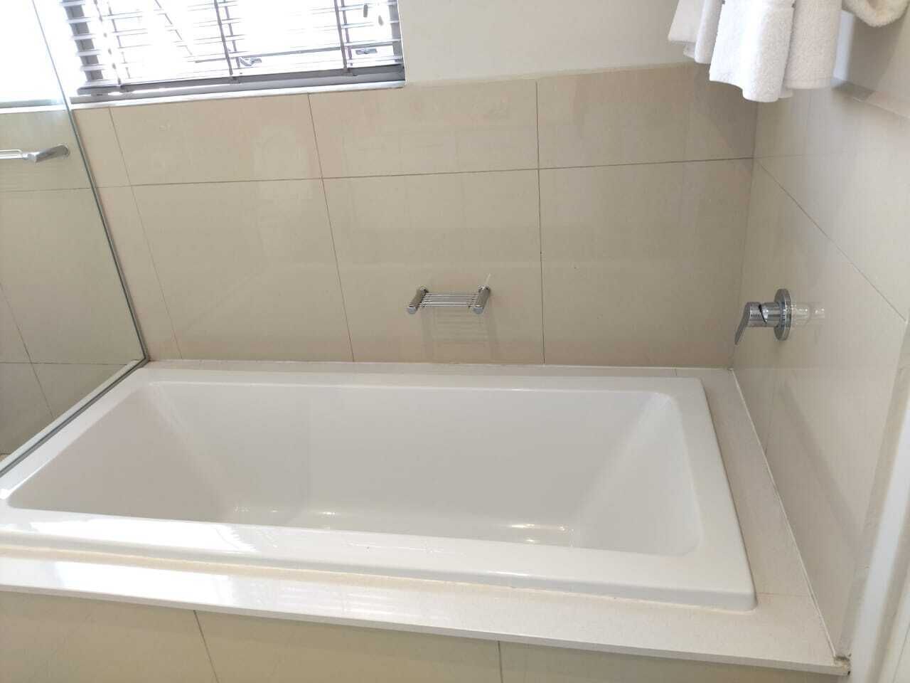 Une baignoire rectangulaire dotée d’un robinet chromé et d’un panneau de commande, installée dans une pièce aux murs recouverts de carreaux beiges et agrémentée d’une fenêtre avec des stores.
