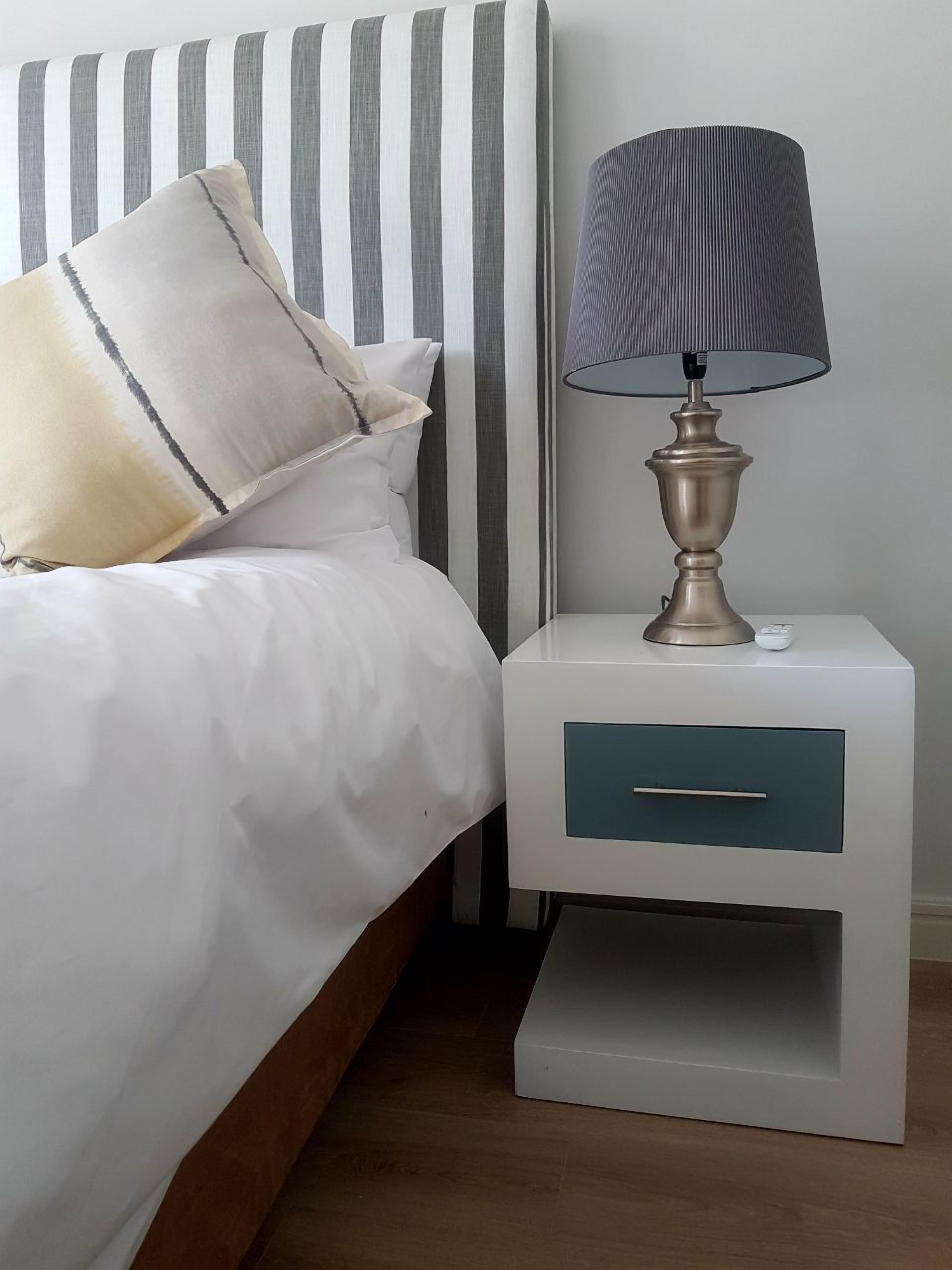 A neatly made bed with white bedding and striped headboard, a bedside lamp, and a nightstand with a drawer.