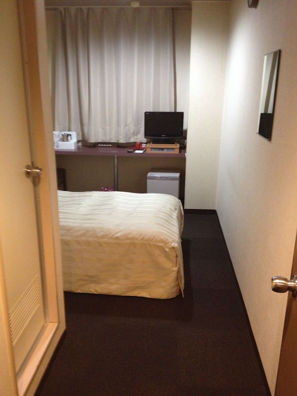Una habitación de hotel estrecha con una cama, un escritorio con una computadora y una pequeña lámpara de escritorio.