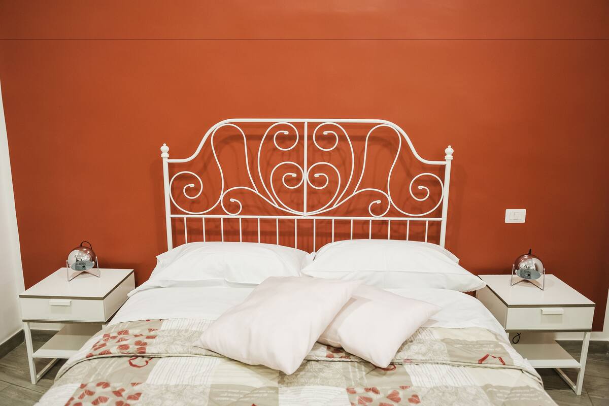 A bedroom with a bed, white nightstands, and a red wall.