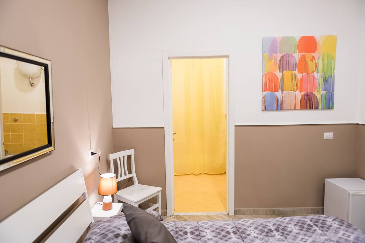 A bedroom with a bed, a chair, a desk, a lamp, a mirror, and a colorful abstract painting on the wall.