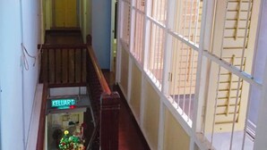 Un couloir avec un escalier en bois, une vue à travers des fenêtres grillagées et un panneau néon indiquant « KELUAR ».