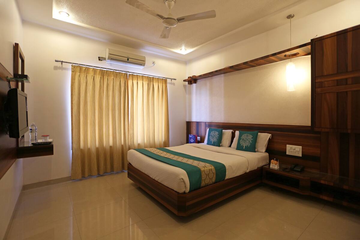 A hotel room with a large bed, a ceiling fan, an air conditioning unit, a bedside table with a lamp, and a wooden headboard.