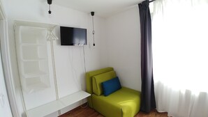 Ein Zimmer mit einem grünen Sofa, einem an der Wand befestigten Flachbildfernseher, einer weißen Bank und einem Fenster mit dunklen Vorhängen.