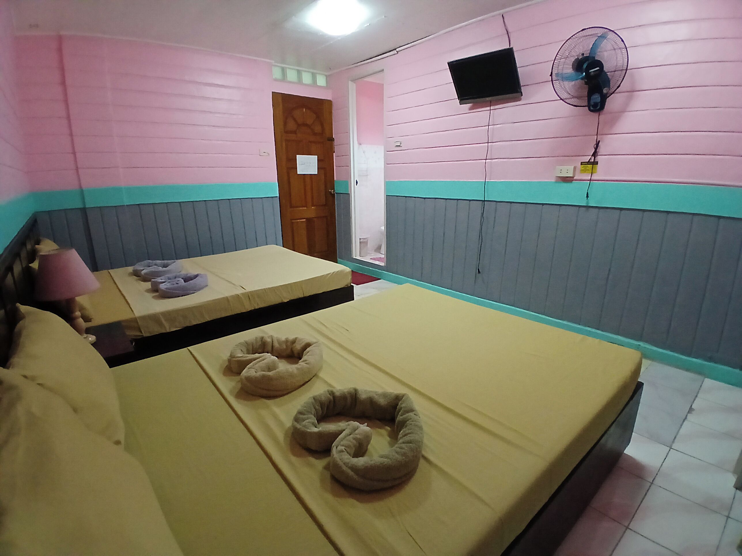 Une chambre avec deux lits, chacun équipé d’un oreiller et d’une couverture. Un ventilateur est fixé au mur, et il y a aussi un téléviseur accroché au mur.