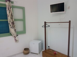 Una habitación con un refrigerador blanco, una mesa de madera con una taza roja, un perchero, una cesta de mimbre y una ventana con contraventanas verdes y cortinas con motivos florales.