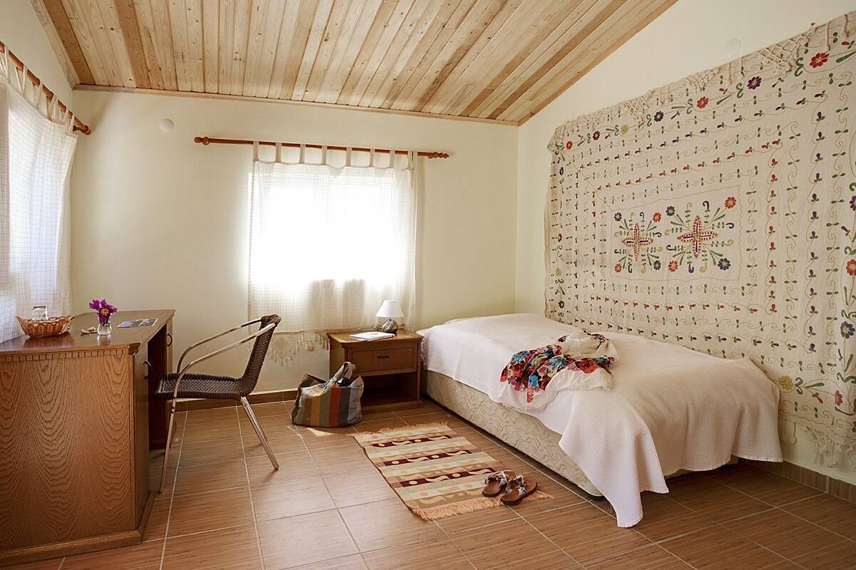 A bedroom with a wooden ceiling, a bed with a floral bedspread, a nightstand with a lamp, a chair, and a small table with a basket.