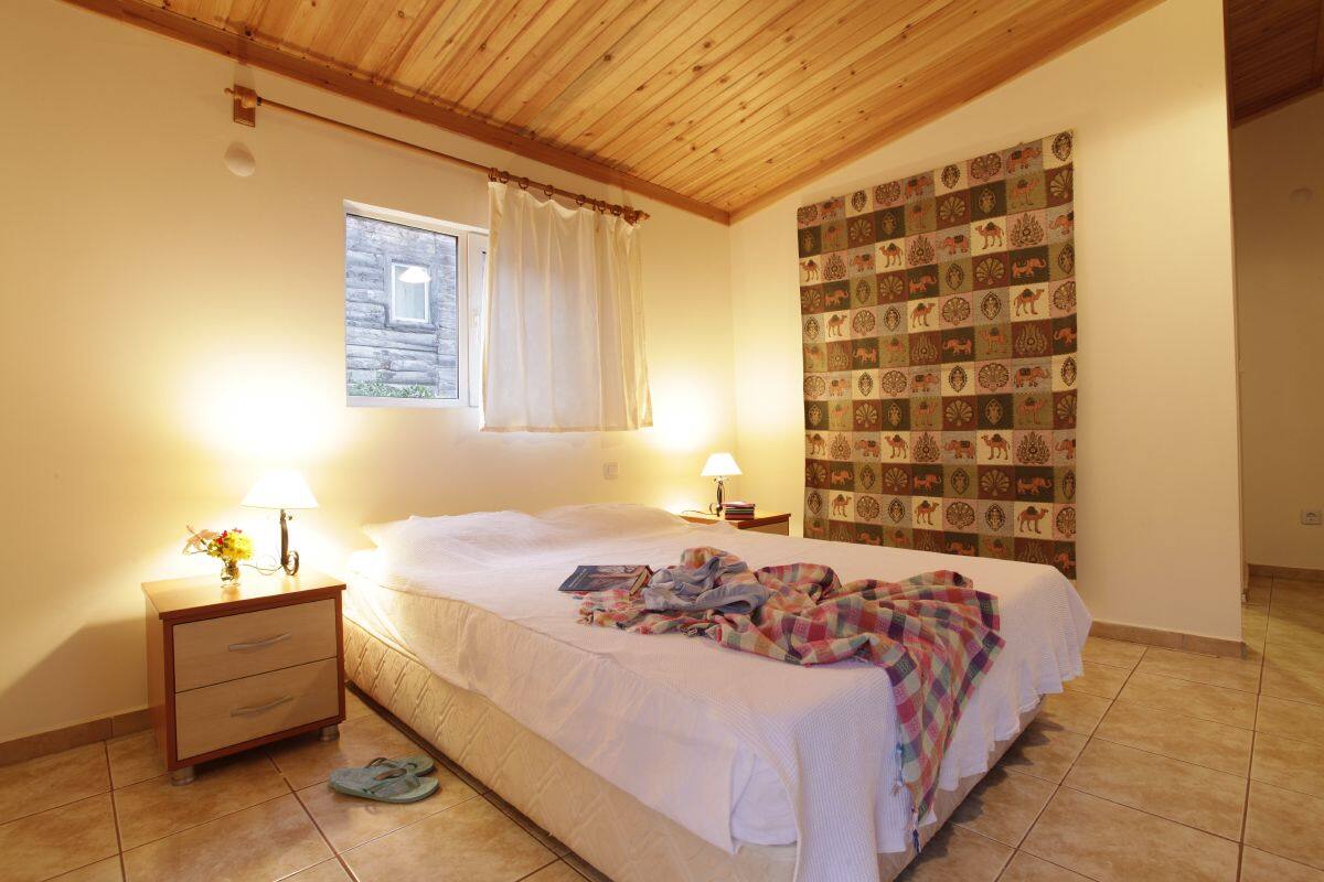A bedroom with a wooden ceiling, a bed with a white bedspread, a nightstand with a lamp, and a wall with patterned wallpaper.