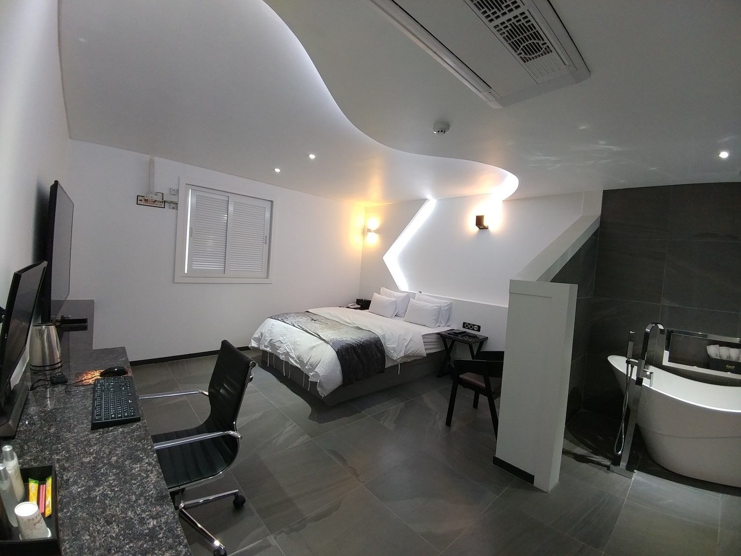 A modern hotel room with a bed, desk, chair, and a bathroom with a bathtub.