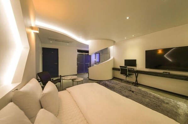 Una habitación de hotel moderna con techo curvo, una cama grande, un televisor de pantalla plana, un escritorio y una zona de estar con una silla y una mesita.