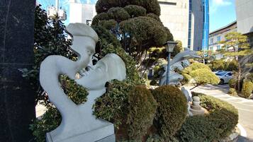 A sculpture of a man and woman embracing, surrounded by well-maintained bushes and trees, with a modern building in the background.