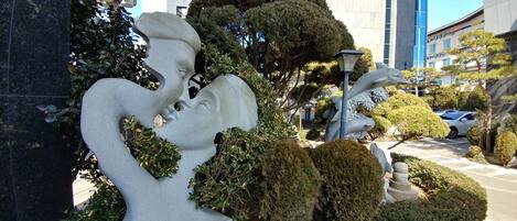 A sculpture of a man and woman embracing, surrounded by well-maintained bushes and trees, with a modern building in the background.