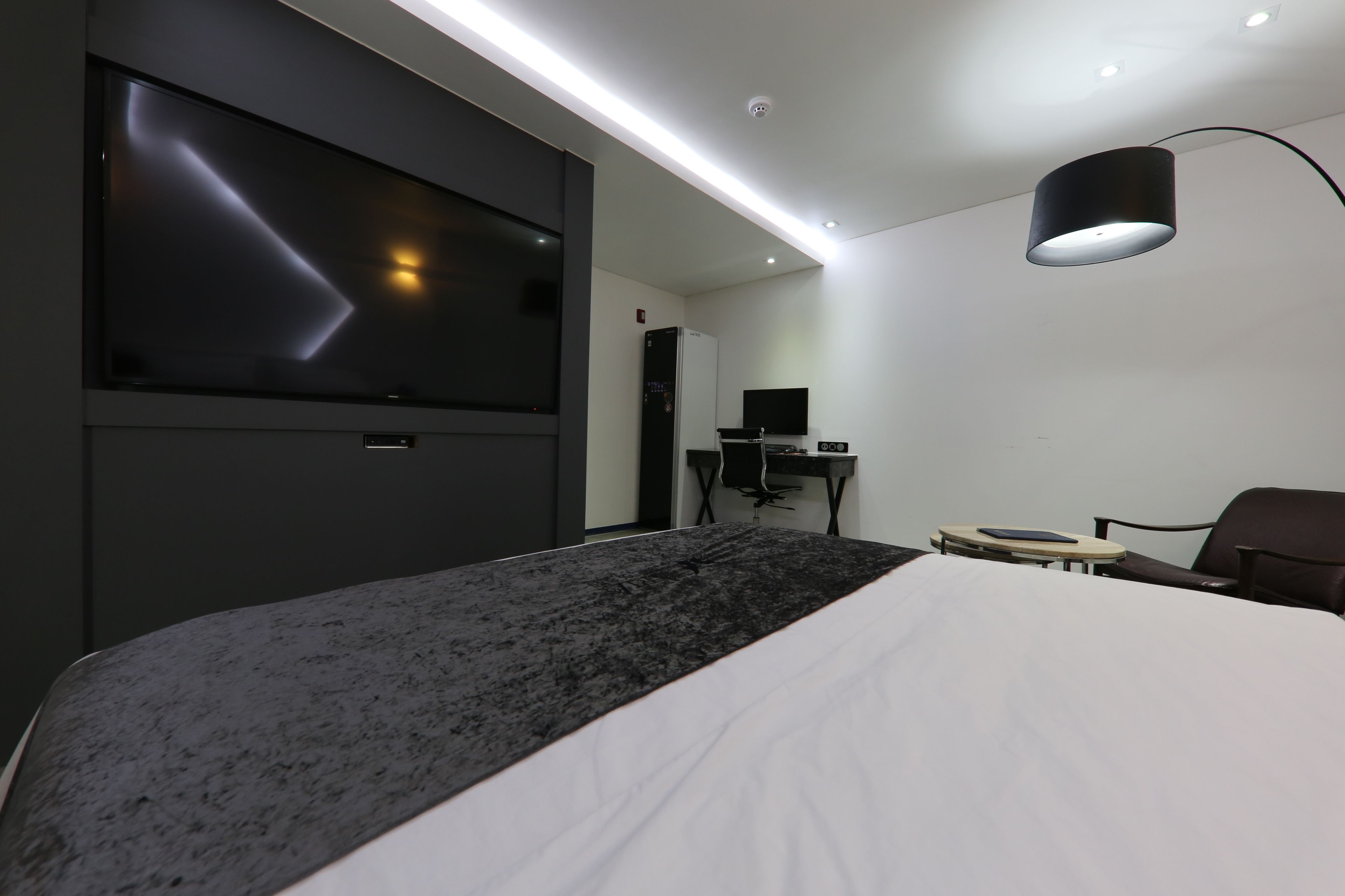 A modern hotel room with a large bed, a flat-screen TV mounted on the wall, a desk with a chair, and a desk lamp.
