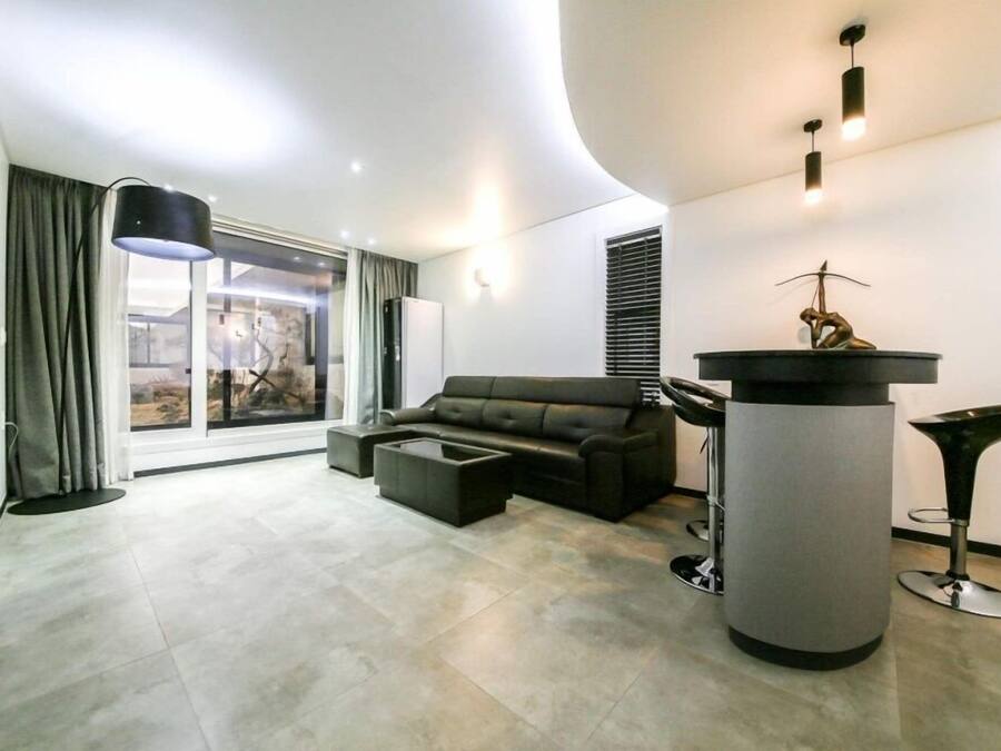 A modern living room with a sectional sofa, a coffee table, a tall column with a sculpture, and a large window with curtains.