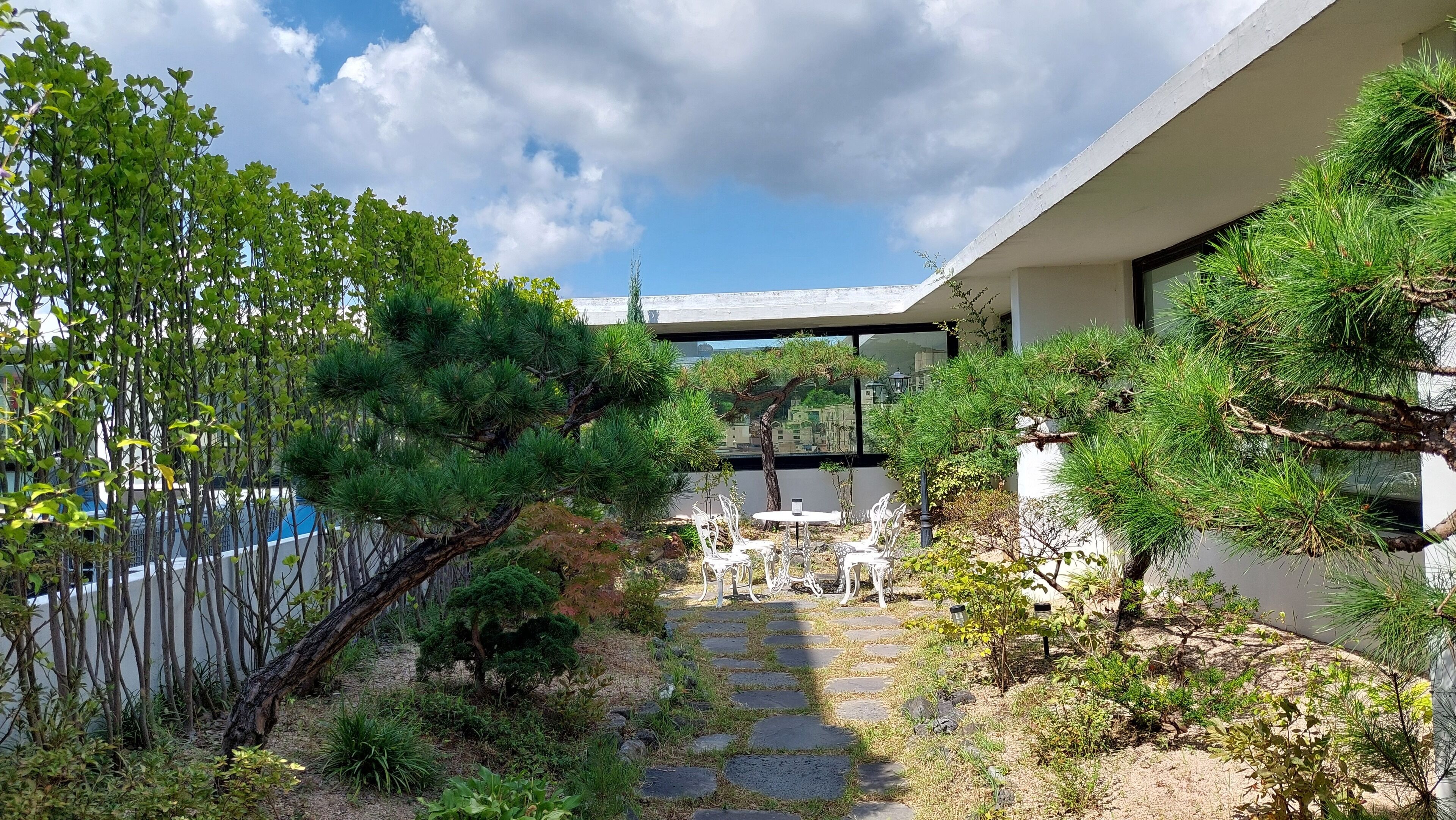 Eine moderne Außenpatio mit Steinböden, umgeben von verschiedenen Pflanzen und Bäumen, und einem Glasgeländer mit Blick auf eine Stadtlandschaft.