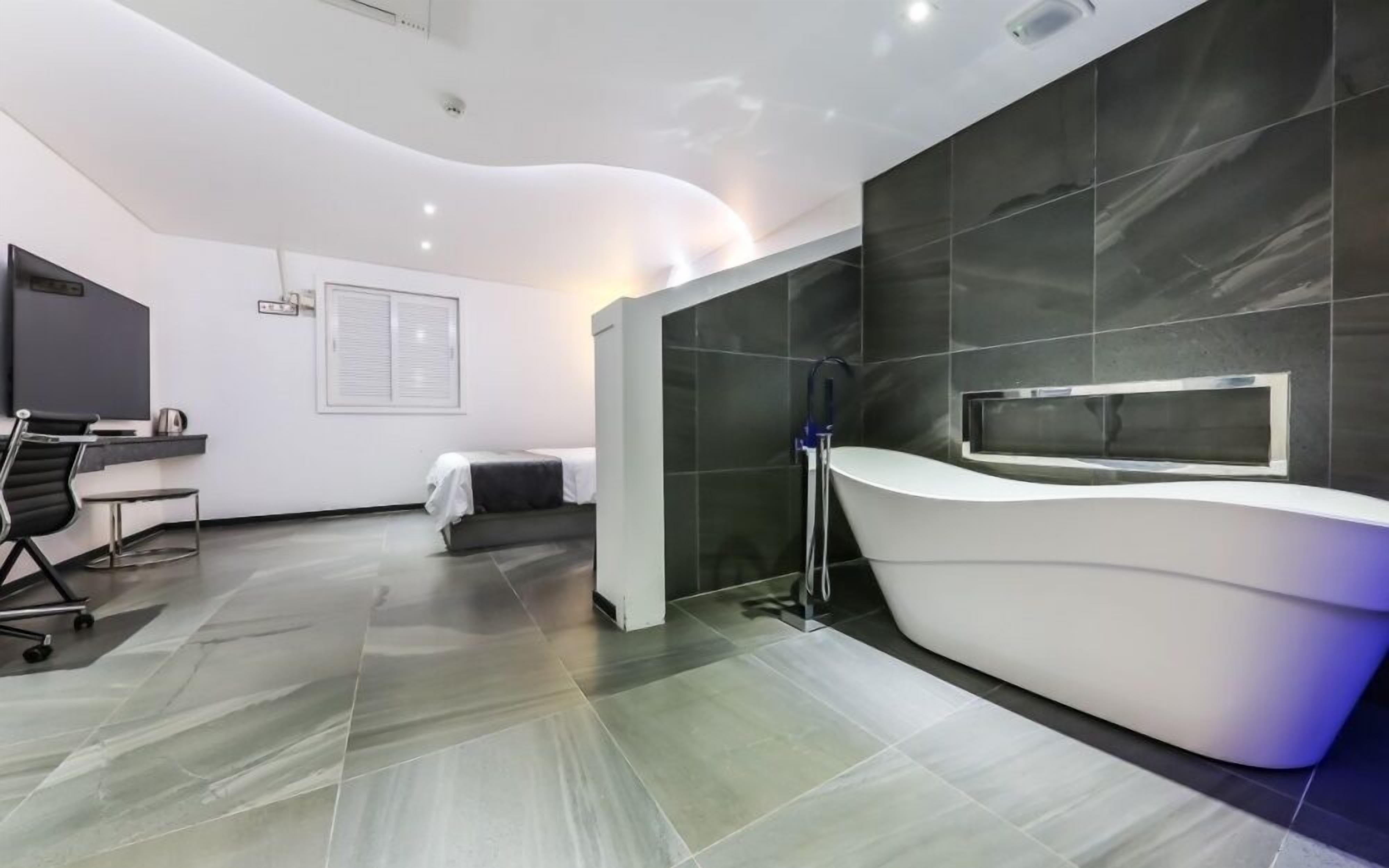 A modern hotel room with a curved reception desk, a bed, a desk with a chair, and a large mirror on the wall.