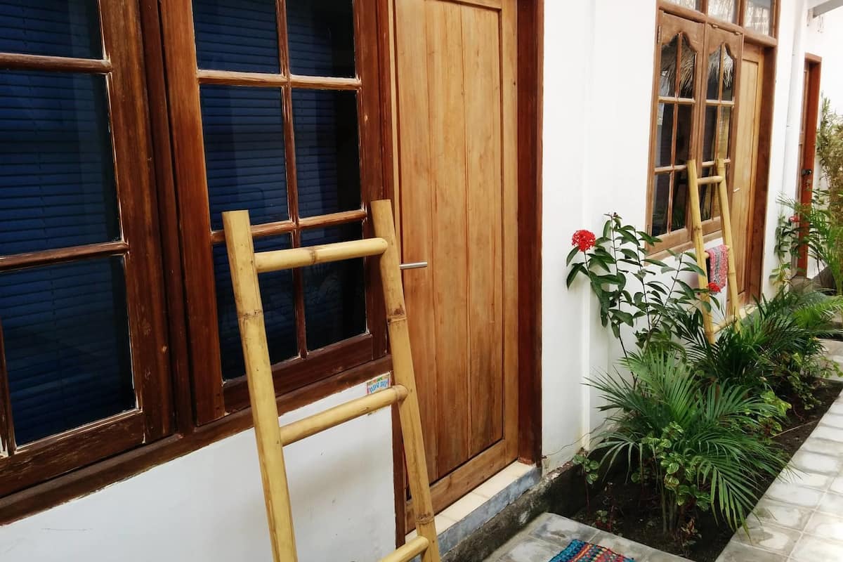 Uma porta de madeira com uma janela à esquerda, uma escada de madeira encostada e um pequeno jardim com plantas e uma flor vermelha.